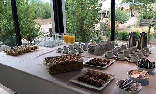 Mesa de coffee break junto a los ventanales, con jugos, termos, tazas y bocados dulces y salados