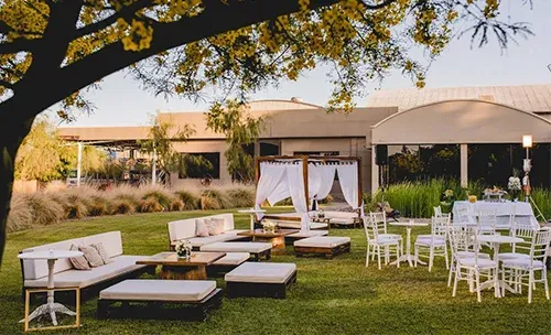 Jardín preparado para una fiesta, con living blanco, mesas redondas y una carpa de telas al atardecer